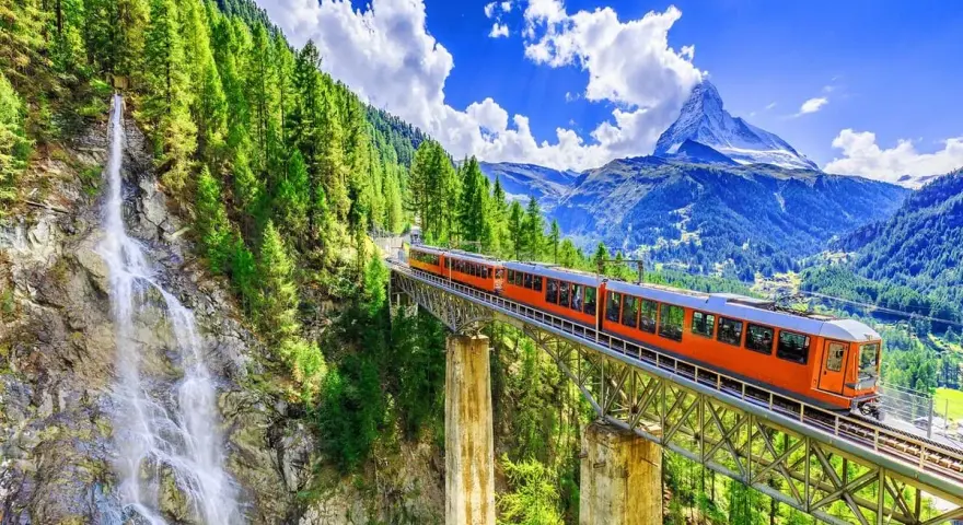 An orange train crosses a bridge over a waterfall, on its way from Zurich to Andermatt. Lush green trees and mountains surround the scenic route.