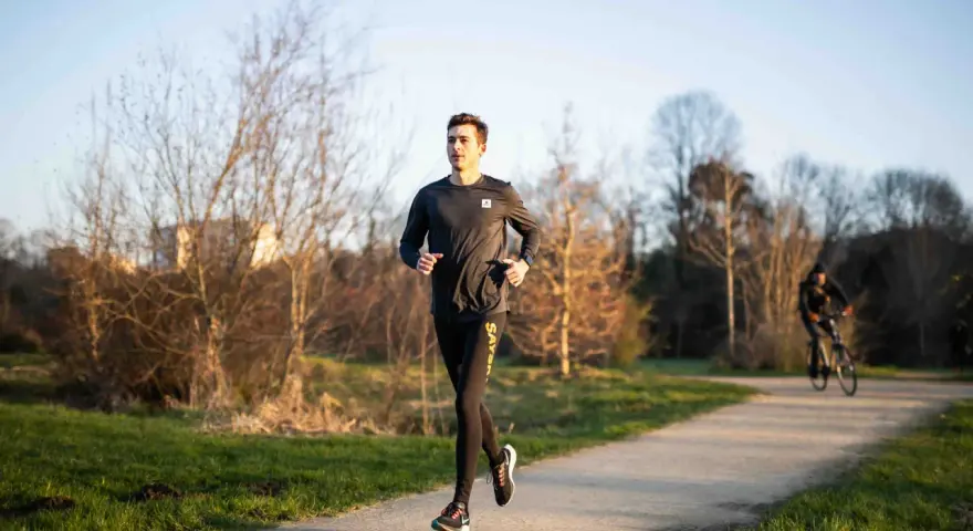 Un jeune homme s'entraîne à la course à pied sur un chemin ensoleillé, entouré d'arbres dénudés. Un cycliste passe en arrière-plan.