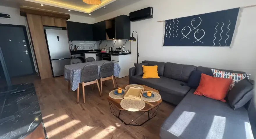 Nowoczesny salon z aneksem kuchennym. Wygodna kanapa, stolik kawowy, stół z krzesłami. W tle lodówka i sprzęty kuchenne. Idealne miejsce na relaks, z myślą o komforcie gości.