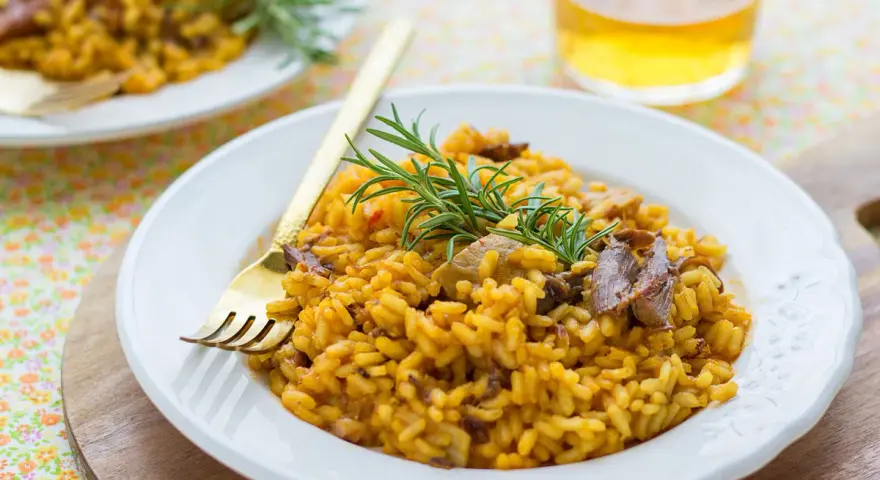 Delicioso arroz con setas y carne, servido en plato blanco con tenedor dorado y ramita de romero.