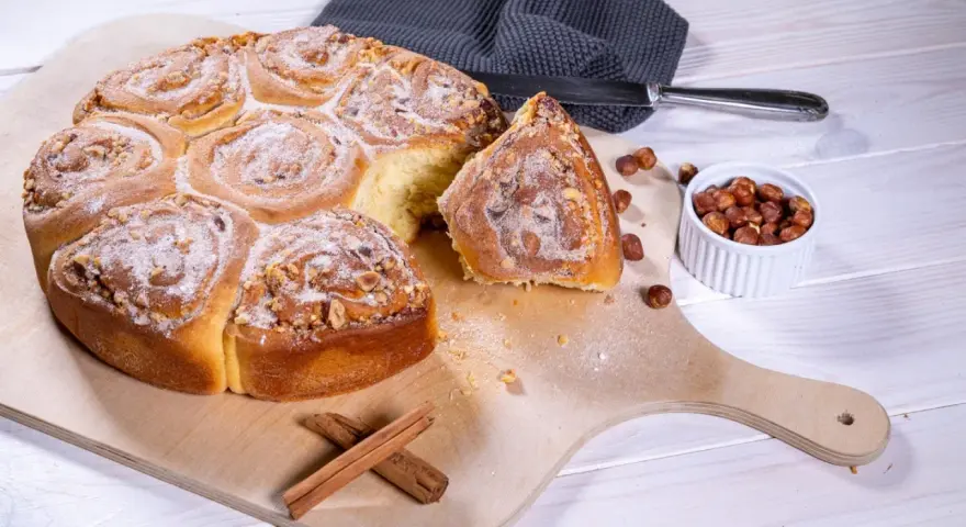 Ein köstliches Zimtschnecken Rezept: goldbraune, mit Nüssen und Puderzucker bestreute Schnecken auf einem Holzbrett, daneben Zimtstangen und Haselnüsse.