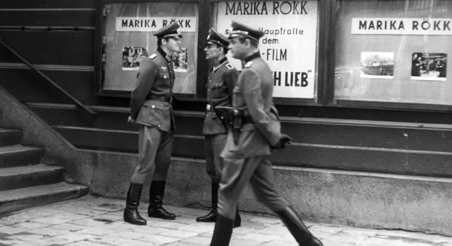 Trzej żołnierze w mundurach stoją przed kinem z plakatem Mariki Rökk. Stawka większa niż życie, teatr historii.
