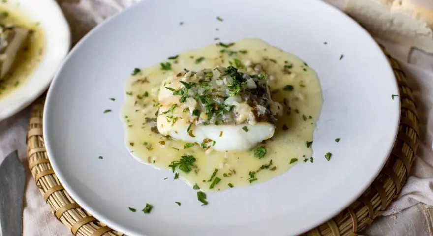 Un delicioso bacalao en salsa verde, con perejil fresco picado y trozos de pan al lado.
