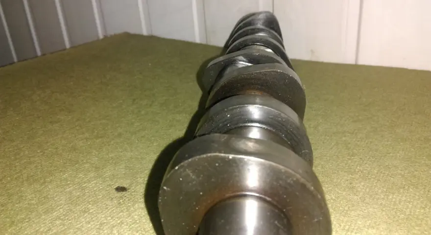 Metalowy, wytarty wałek rozrządu leży na zielonym materiale. Widać ślady zużycia na jego krzywkach.
