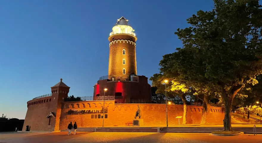 Wieczorny spacer przy latarni morskiej Kołobrzeg. Oświetlona ceglana budowla z czerwonymi akcentami i drzewa tworzą magiczny klimat.