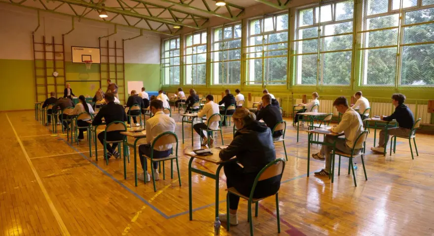 Uczniowie siedzą przy ławkach w sali gimnastycznej, pisząc egzamin.