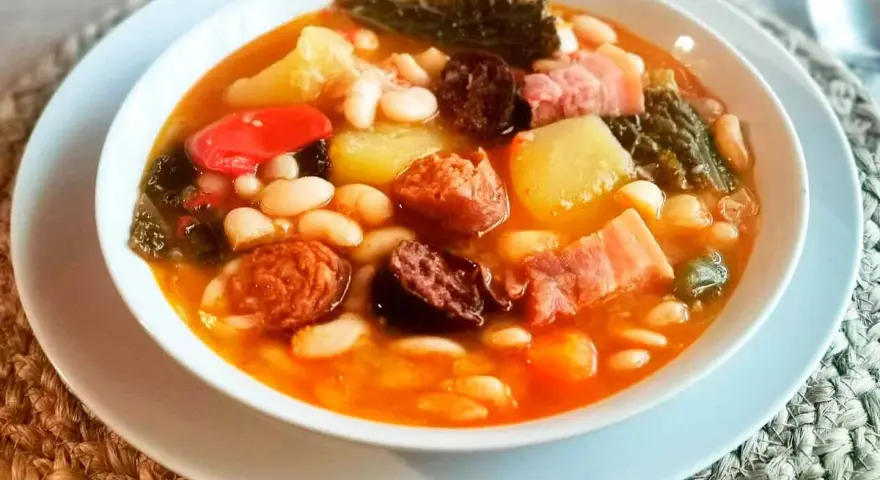 Un reconfortante cocido montañés, rebosante de alubias blancas, patatas, chorizo, morcilla y tocino, servido en un cuenco blanco.