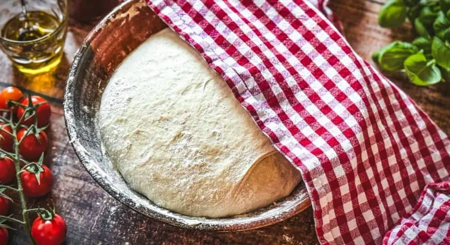 Frischer Pizzateig ruht in einer Schüssel, bedeckt mit einem rot-weiß karierten Tuch. Zutaten für ein Pizzateig Rezept wie Tomaten und Öl liegen bereit.