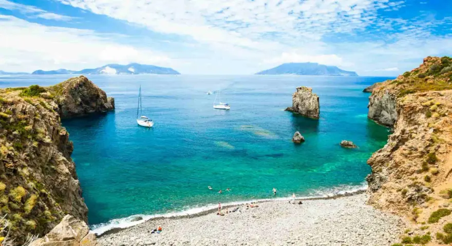 Rocky coastline with a pebble beach, turquoise water, sailboats, and distant islands under a blue, cloudy sky.