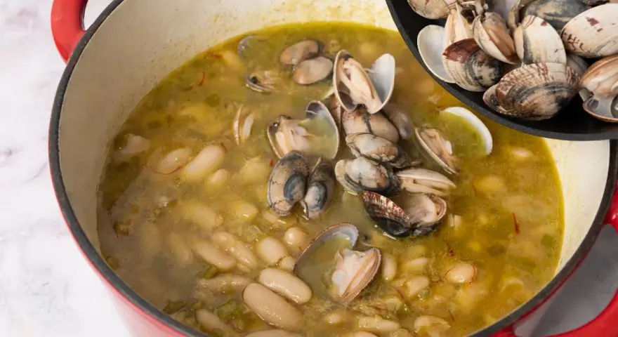 Alubias cremosas con almejas en una olla roja, añadiendo más almejas de una sartén.