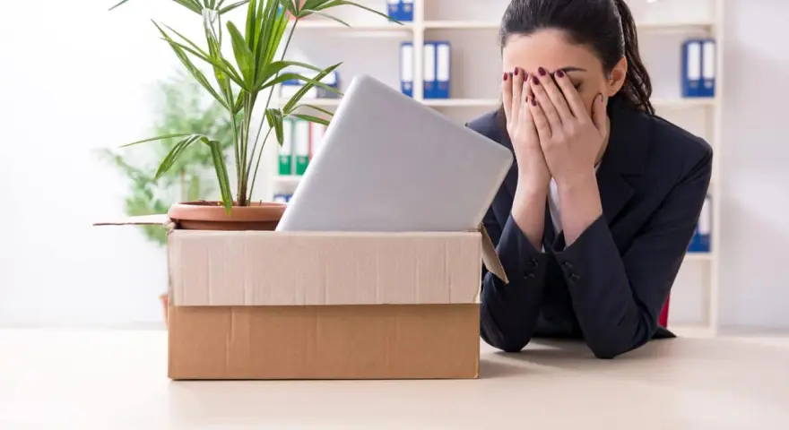 Verzweifelte Frau mit Karton und Laptop im Büro. Sie muss eine innerbetriebliche Versetzung ablehnen und ist sichtlich mitgenommen.