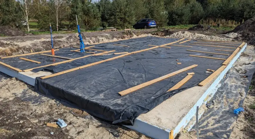 Gotowy fundament pod dom szkieletowy, z czarną folią i drewnianymi belkami, czeka na dalsze prace budowlane.