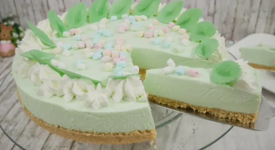 Eine helle, cremige Philadelphia Torte Waldmeister mit bunten Marshmallows und grünen Blättern verziert. Ein Stück wird gerade herausgeschnitten.