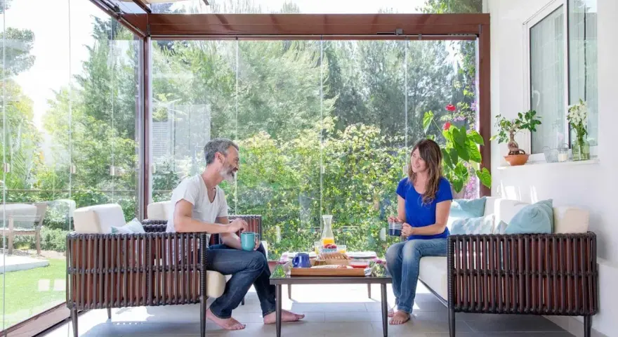 Pareja disfrutando de un café en un **porche acristalado** con vistas a un jardín exuberante.