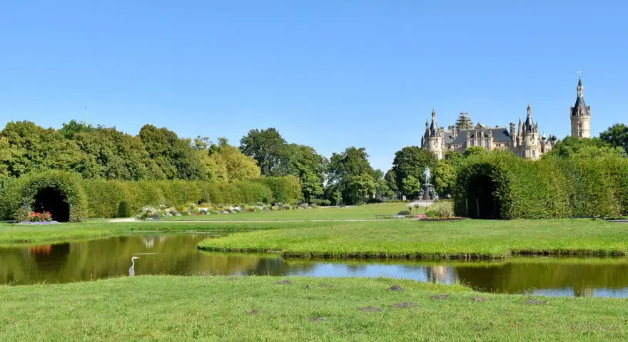 Ein Reiher steht am Ufer eines Teichs im Schweriner Schlossgarten. Im Hintergrund thront das Schloss über grünen Bäumen.
