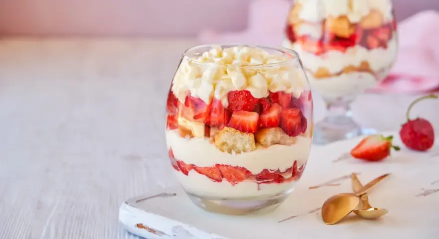 Postre en vaso con capas de crema, fresas y bizcocho, cubierto con trozos de chocolate blanco.