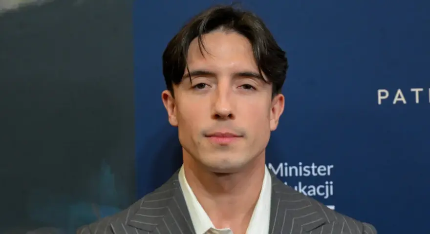 Dawid Kwiatkowski w szarym garniturze na tle niebieskiej ściany z napisami.