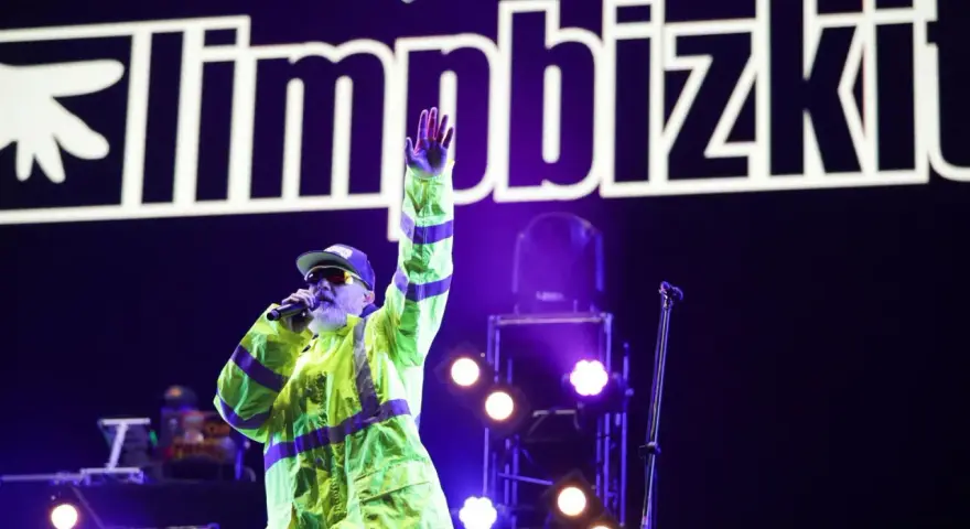 Fred Durst z Limp Bizkit na koncercie, w neonowej kurtce, z ręką w górze. Tło to logo zespołu.