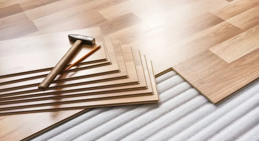 Tablas de suelo laminado apiladas con martillo y lápiz, listas para aislar suelo frío sobre una base acolchada.