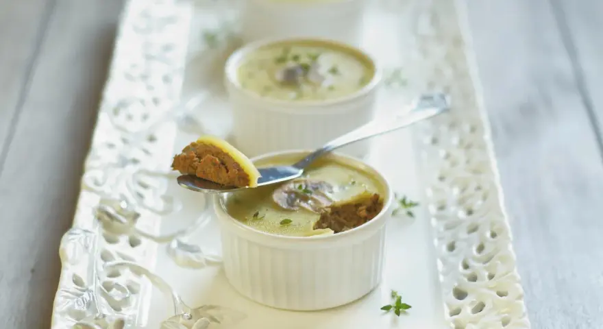 Trois ramequins de foie de volaille, ornés de champignons et de persil, sur un plateau ajouré. Une cuillère présente une bouchée de ce délice.