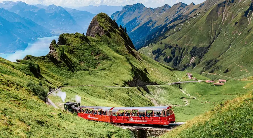 A red Brienz Switzerland train winds through lush green mountains, with a stunning blue lake in the distance.