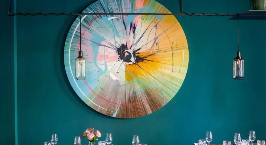 Ein bunter, runder Kunst-Splash ziert die Wand einer stilvollen Werkstatt in Berlin. Tische sind gedeckt, bereit für Gäste.