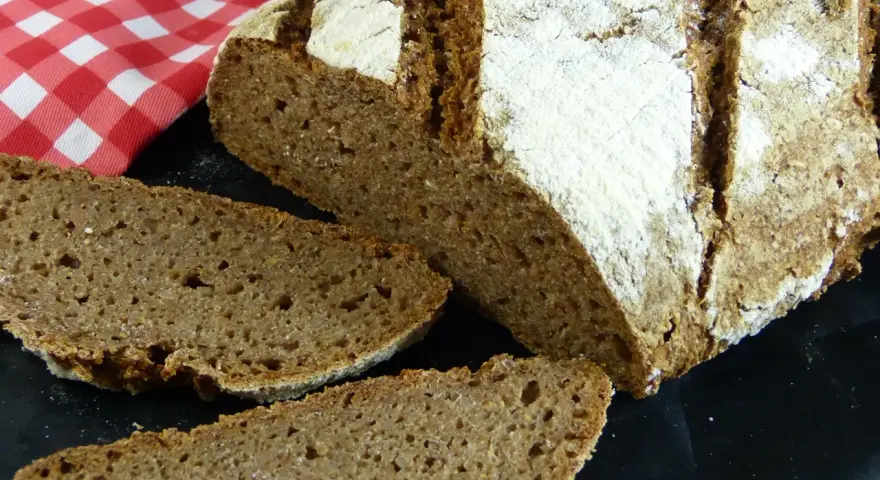 Ein rustikales tschechisches Brot, frisch aufgeschnitten, mit einer kräftigen Kruste und einer saftigen Krume. Daneben liegt eine rot-weiß karierte Serviette.