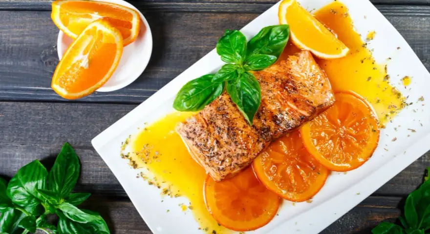 Delicioso salmón a la naranja, servido con rodajas de naranja y hojas de albahaca fresca.