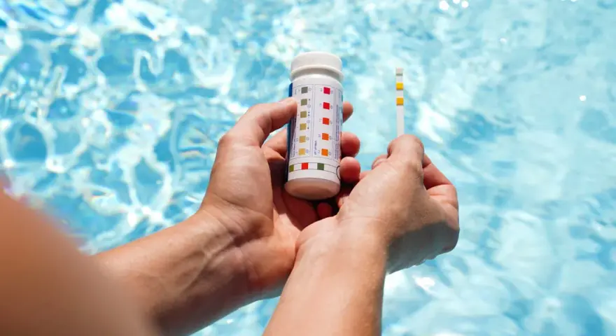 Hands hold a pool test strip and bottle, showing how to make pool water clear by checking chemical levels.
