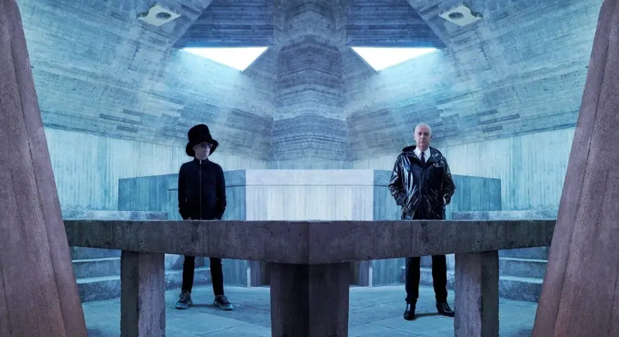 Pet Shop Boys in futuristischer Betonarchitektur. Neil Tennant mit Zylinder, Chris Lowe im glänzenden Mantel.