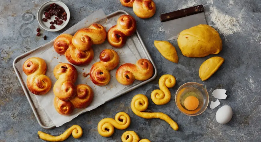 En bricka med gyllene, nybakade lussekatterna, redo att avnjutas. Bredvid ligger ingredienser som russin, deg och ett ägg. Dessa är de godaste lussekatterna!