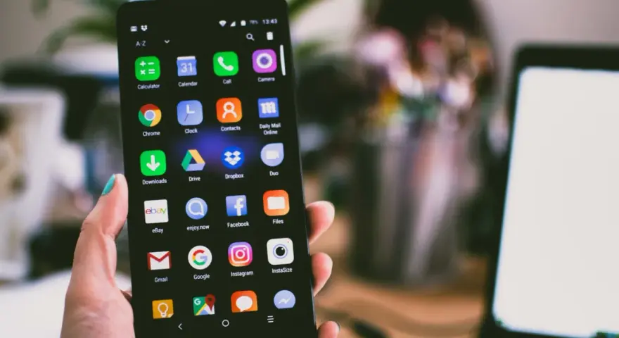 Dłoń trzyma smartfon z widocznym ekranem pełnym ikon aplikacji, w tym ikoną wiadomości SMS.