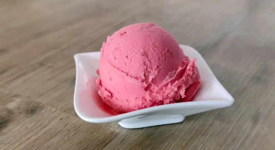 Ein Löffel cremiges Himbeersorbet in einer weißen Schale auf Holzhintergrund.