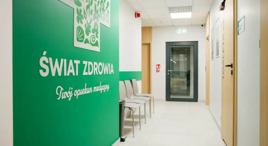 Zielona ściana z napisem "Świat Zdrowia" i logo, korytarz kliniki medycznej z drzwiami i krzesłami.