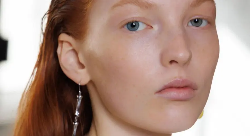 Primo piano di una giovane donna con capelli rossi e occhi azzurri, un neo della bellezza sul viso. Indossa un orecchino pendente.