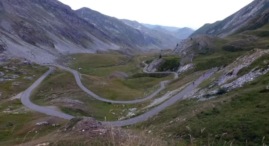Strada tortuosa che sale verso il colle dei morti, tra montagne verdi e rocciose. Due ciclisti affrontano le curve.