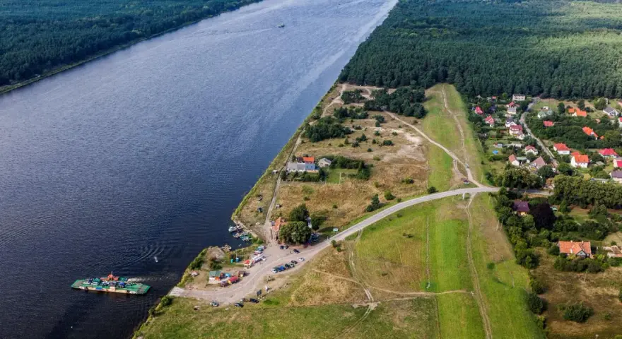 Widok na Wisłę i jej ujście. Prom przewozi samochody, a w oddali widać las i zabudowania.