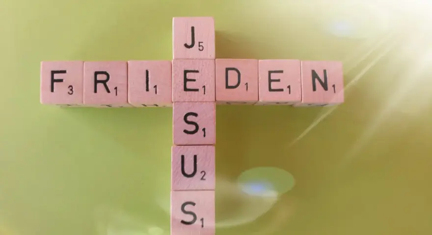 Buchstabensteine bilden ein Kreuz mit den Wörtern "FRIEDEN" und "JESUS". Ein Zitat aus Johannes 14.