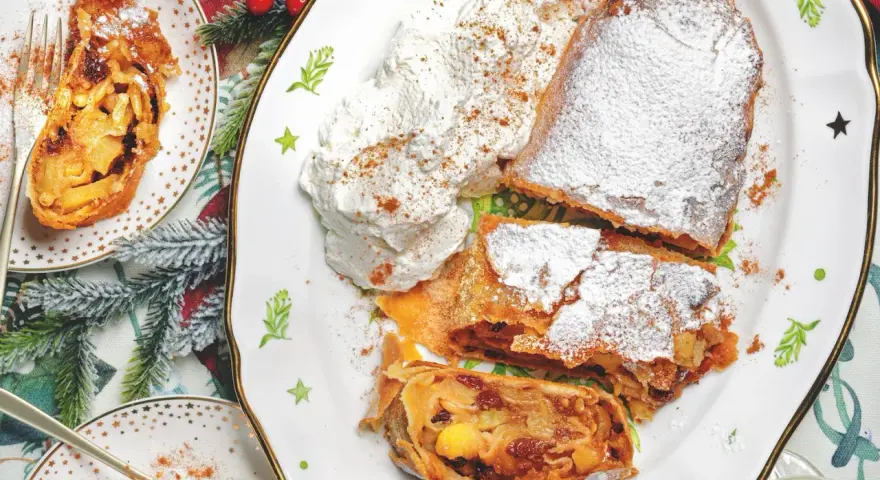 Delizioso strudel di mele con pasta sfoglia, cosparso di zucchero a velo e servito con panna montata e cannella.
