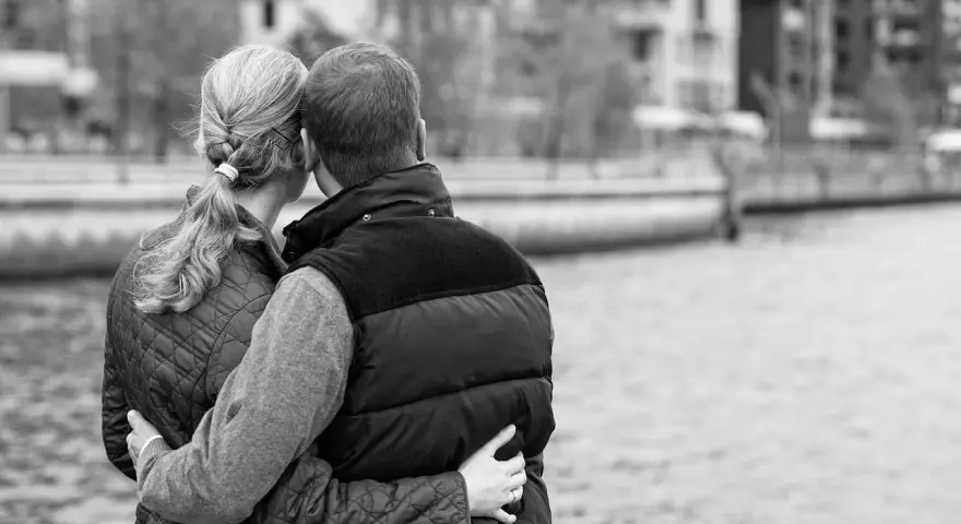 Para przytulająca się nad wodą. Jej ręka obejmuje jego talię, a jego ramiona otulają ją. Delikatny dotyk w relacjach damsko-męskich.