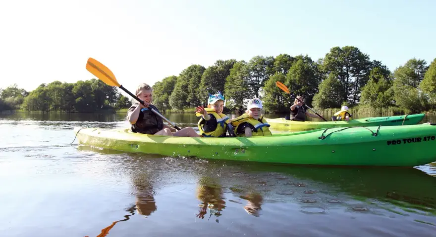 Rodzina pływa kajakami po spokojnej rzece. Dzieci w kamizelkach ratunkowych machają do obiektywu, ciesząc się słonecznym dniem na wodzie.