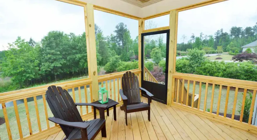 A screened-in porch with two Adirondack chairs, a small table, and a green lantern. The porch offers a view of lush trees and a suburban landscape.