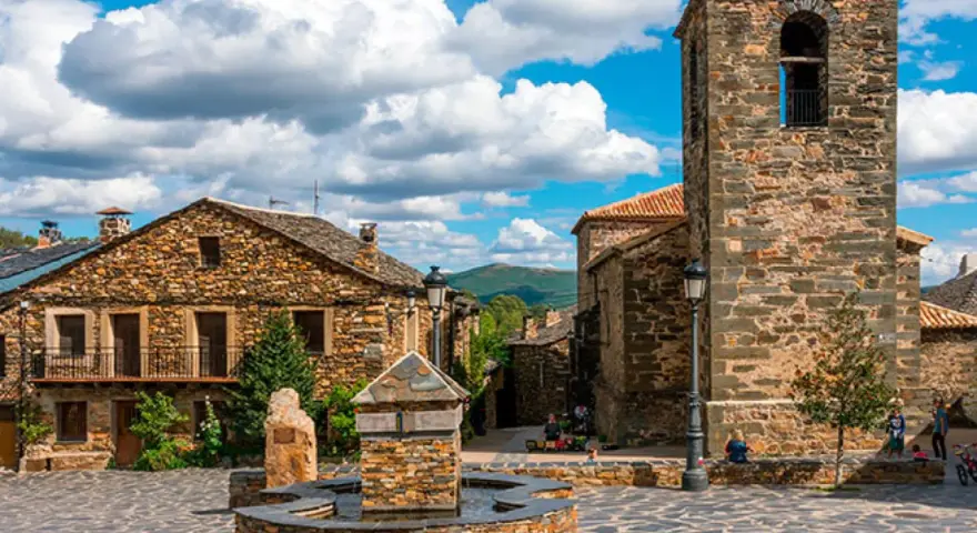 Plaza con fuente de piedra y campanario en un pueblo de la ruta negra Guadalajara. Casas de piedra y cielo azul con nubes.