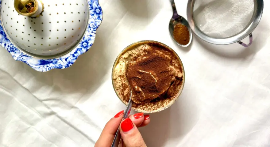 Deliziosa crema al caffè in bicchiere, cosparsa di cacao. Accanto, un elegante contenitore bianco e oro e un cucchiaino con cacao.