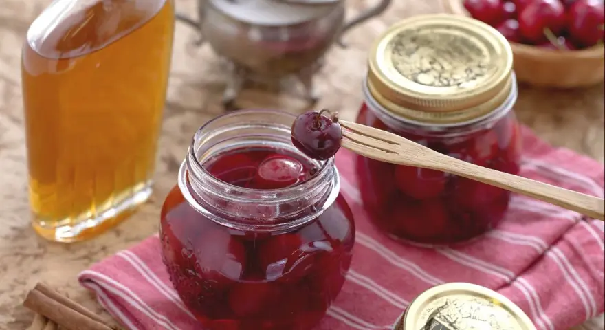Frascos de mermelada de cerezas casera, una botella de licor y canela en rama. Una cereza en un tenedor de madera lista para probar.