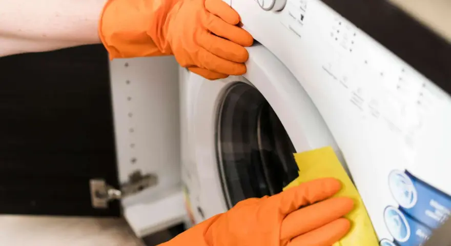 Hände in orangen Handschuhen reinigen die Waschmaschine. Ein gelber Lappen wischt über die Oberfläche, um sie sauber zu halten.