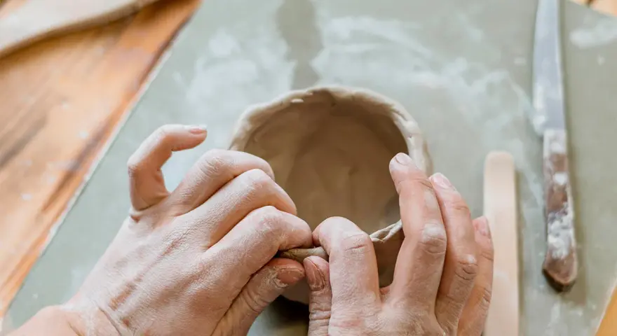 Mani che modellano argilla per creare una ciotola. Utensili da ceramica e un coltello sono visibili. Un ottimo punto di partenza per chi cerca dove trovare l'argilla.
