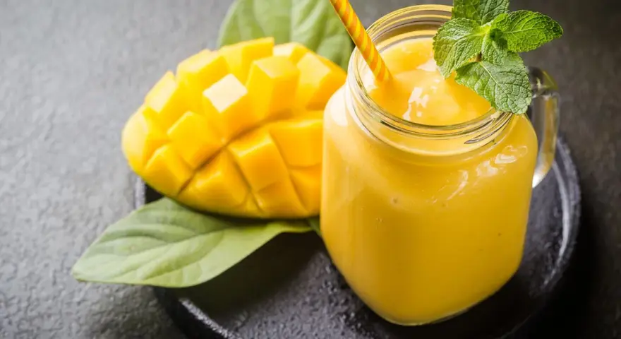 Un smoothie mangue onctueux dans un pot en verre, garni de menthe fraîche, à côté d'une moitié de mangue coupée en dés.