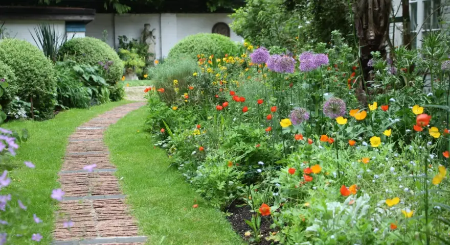 Ein geschwungener Weg aus Ziegelsteinen schlängelt sich durch einen üppigen bauerngarten mit leuchtenden Blumen und grünem Rasen.