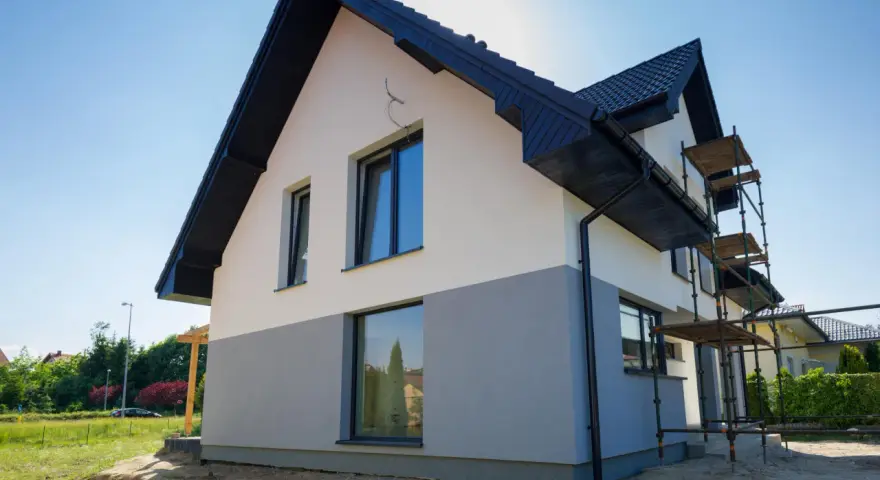 Nowy dom w trakcie prac wykończeniowych. Elewacja dwukolorowa, biało-szara, z czarnym dachem i rynnami. Widoczne rusztowania.
